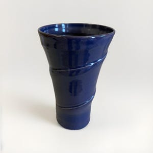 May include: A glossy, dark blue ceramic vase with a flared top and a gently curved, spiral design. The vase has a smooth, reflective surface and a wide opening, suitable for displaying flowers or as a decorative piece.