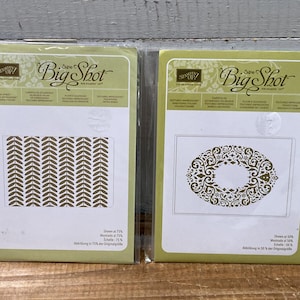 Può includere: Due confezioni verdi e bianche di fustellatrici e goffratrici Stampin' Up! Big Shot. Una confezione presenta un motivo a frecce ripetute, l'altra ha un design floreale ovale.