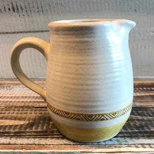 May include: Cream-colored ceramic pitcher with a curved handle and a spout. The pitcher has a yellow band at the base and a decorative geometric pattern in a band above the yellow. The pitcher is on a wooden surface.