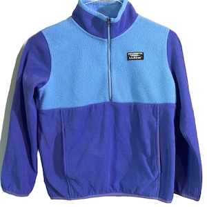 May include: A blue and purple fleece pullover with a quarter-zip closure. The top portion is light blue, while the lower section and sleeves are purple. The brand logo is visible on the chest. This warm fleece is perfect for outdoor activities.