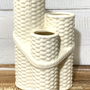Puede incluir: Jarrón de cerámica blanquecina con textura de cesta tejida. El jarrón tiene tres aberturas cilíndricas de diferentes tamaños, conectadas por una banda curva y texturizada. Un elemento decorativo para el hogar.