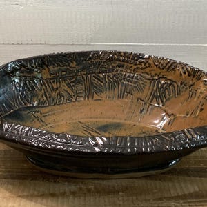 Op de afbeelding: Handgemaakte keramische kom met een donkerbruine buitenkant en een lichtere bruine binnenkant. De kom heeft een getextureerde rand en een uniek patroon aan de binnenkant. Een decoratief stuk voor in huis.