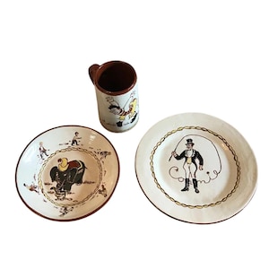 May include: A ceramic circus-themed set featuring a mug and two plates. The mug has a brown handle and a white body with cartoon figures. The plates have circus illustrations with brown rims. One plate shows an elephant, and the other a ringmaster.