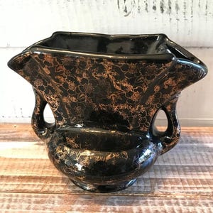 Pode incluir: Vaso de cerâmica preta com borda alargada e duas alças. O vaso é decorado com um padrão floral dourado. O vaso tem um acabamento brilhante e assenta numa base pequena e redonda. O vaso é provavelmente vintage.