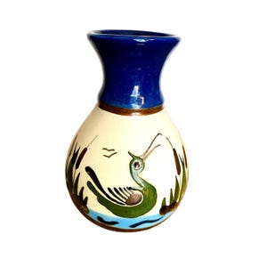 May include: A ceramic vase with a blue rim and a cream-colored body. The vase features a hand-painted design of a green bird, reeds, and water. The vase is approximately 6 inches tall.