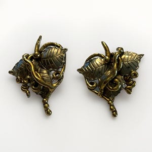 May include: A pair of vintage gold-toned leaf-shaped clip-on earrings. Each earring features detailed leaf and vine designs with a textured finish. The earrings have a classic, elegant style.