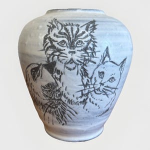 以下が含まれることがあります： 丸みを帯びた形状の白いセラミック花瓶。黒インクで手描きされた3匹の猫のイラストが特徴です。花瓶はわずかにテクスチャ加工された表面と暗い縁を持ち、気まぐれで芸術的なデザインを示しています。