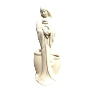 Puede incluir: Figura de cerámica color crema de la Virgen María sosteniendo a un bebé. La estatua tiene una base en forma de jarrón con dos aberturas. La figura lleva una túnica con capucha.