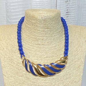 May include: A vintage necklace featuring a strand of blue beads and a gold-toned pendant with blue enamel accents. The pendant has a curved, layered design. The necklace is displayed on a mannequin.