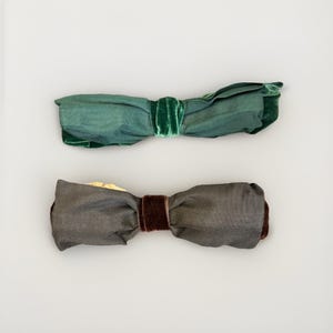 May include: Two fabric bow ties. The top bow tie is green with a green velvet center. The bottom bow tie is gray with a brown velvet center. Both bow ties are on a white background.