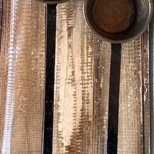 May include: Two antique metal ladles with long, black handles and circular bowls. The bowls are a dark, tarnished metal, and the handles have a loop at the end. The ladles are displayed against a weathered wooden background.