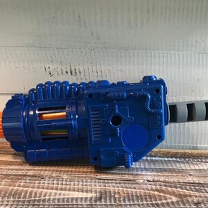 Op de afbeelding: Een blauw en oranje speelgoedpistool met een grijze handgreep. Het speelgoedpistool heeft een futuristisch ontwerp met meerdere details, waaronder een geribbelde bovenkant en een reeks rechthoekige openingen. De handgreep is cilindrisch met een gegroefde grip.