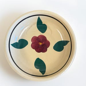 May include: A round, cream-colored ceramic plate with a hand-painted floral design. The center features a burgundy flower with a yellow center, surrounded by four green leaves. A black line borders the rim.