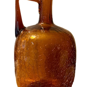 Puede incluir: Jarra de vidrio color ámbar con asa. La jarra tiene un cuerpo redondeado y un cuello largo y estrecho. La superficie del vidrio tiene una textura agrietada, lo que añade interés visual. Esta jarra decorativa es adecuada para exhibir o como jarrón.