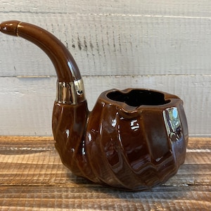 Könnte beinhalten: Ein brauner, pfeifenförmiger Aschenbecher aus Keramik mit glänzendem Finish. Der Aschenbecher hat einen gebogenen Stiel und einen schalenförmigen Körper mit einem strukturierten, gerippten Design. Der Stiel hat ein goldfarbenes Band in der Nähe der Schale.