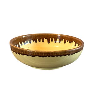 Puede incluir: Un cuenco de cerámica redondo con exterior color crema e interior amarillo cálido. El borde presenta un diseño de esmalte marrón goteado. El cuenco parece hecho a mano.