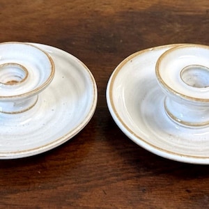 May include: A pair of matching ceramic candle holders featuring a circular base, a central raised cup for a candle, and a small side handle. The pieces are finished in a glossy off-white glaze with visible brown trim along the edges and rims.