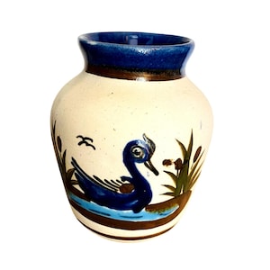 May include: A ceramic vase with a cream-colored body, a blue rim, and a brown band. The vase features a hand-painted design of a blue swan on a blue water line, with brown and green reeds and two small birds.