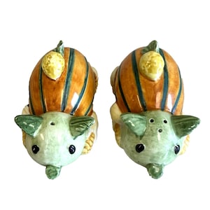 May include: A pair of ceramic salt and pepper shakers. Each shaker is shaped like a pig with a pumpkin on its back. The pumpkins are orange with green stripes and a yellow lemon on top. The pigs are light green with black eyes.
