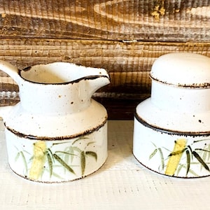 May include: A white ceramic creamer and sugar bowl set. The set features a speckled finish with a brown rim and a bamboo design in green and yellow. The creamer has a handle and spout.