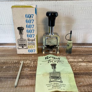 May include: A vintage Regal 607 six-wheel numbering machine with a black handle, a box, and an instruction sheet. The box and instruction sheet have the number 607 printed on them. Also included is a small bottle of oil and a pencil.