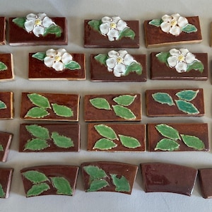 May include: A collection of brown ceramic tiles with white flower and green leaf designs. Some tiles feature a complete flower, while others display leaves. The tiles are rectangular and have a glossy finish.
