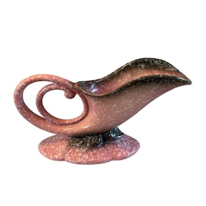 May include: A vintage ceramic gravy boat in a speckled pink and black color scheme. The boat features a distinctive handle with a looped design and a flared spout. The base is a scalloped shape.