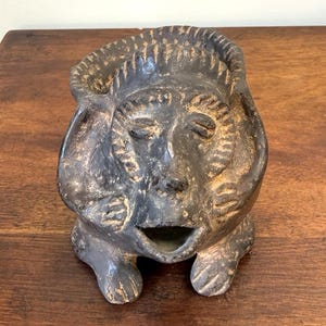 Op de afbeelding: Een donkere, verweerde keramische sculptuur van een gestileerd gezicht met gesloten ogen en een open mond. De sculptuur heeft een getextureerd oppervlak en wordt ondersteund door kleine voetjes. Het stuk is waarschijnlijk een kunstvoorwerp of een decoratief item.