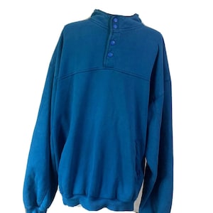 Puede incluir: Una sudadera azul turquesa con cuello alto y tres botones. La parte superior de manga larga tiene un corte holgado y un diseño de color liso. La prenda está hecha de un material suave y es adecuada para ropa casual.