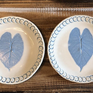 May include: Two ceramic plates with a cream-colored base and a blue leaf design in the center. The rims are decorated with a repeating blue pattern. The plates are displayed on a wooden surface.