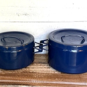 May include: Two navy blue enamel pots with lids and handles. The pots have a round shape and are sitting on a wooden surface. The handles are black and angular. The background is a white wooden wall.