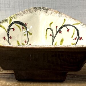Pode incluir: Uma tigela de servir de cerâmica marrom com uma parte superior triangular e uma base retangular. O interior é creme com um desenho floral preto e verde e detalhes em vermelho. A borda tem um padrão salpicado e texturizado.