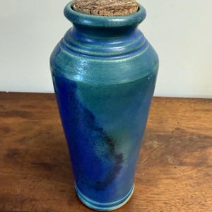 Puede incluir: Un tarro de cerámica alto con cuerpo cilíndrico cónico y borde acampanado. El exterior presenta un esmalte mezclado de azul cobalto profundo y verde azulado con acentos oscuros moteados. Un tapón de corcho natural texturizado cierra la abertura.