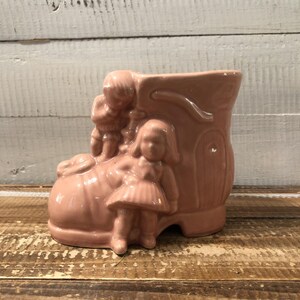 Puede incluir: Una maceta de cerámica rosa en forma de bota con figuras en relieve de niños. La bota tiene una punta redondeada y una parte superior alta, con un diseño decorativo. Los niños visten ropa vintage.
