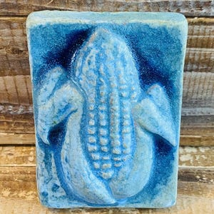 May include: A rectangular, ceramic tile with a relief of a peanut. The tile is primarily light blue with darker blue accents, creating a textured, three-dimensional effect. The peanut design is centered on the tile.