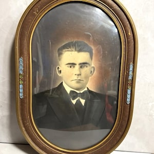 Puede incluir: Retrato antiguo ovalado en un marco marrón con ribete dorado. El retrato presenta a un hombre con traje negro y pajarita. El marco tiene detalles decorativos azules y rojos. El retrato está detrás de un cristal.