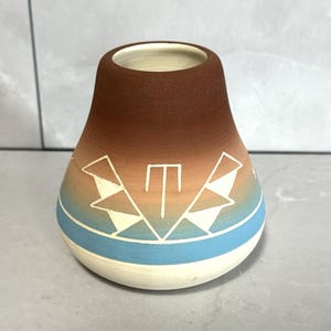 May include: A handcrafted ceramic vase with a rounded base and a narrow opening. The vase features a colour gradient from brown to beige, with a blue stripe and white geometric designs. The vase is approximately 13 cm tall.