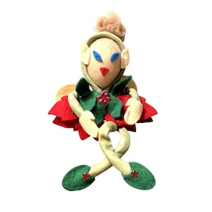 May include: A handmade felt Christmas elf doll with a beige face, blue eyes, and a red nose. The elf wears a green and red outfit with a flower accent. The elf has green shoes with red star accents and beige ears.