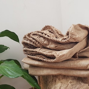 Könnte beinhalten: Ein Stapel brauner Leinen-Spannbettlaken, gefaltet und einsatzbereit. Die Laken sind aus natürlichem Leinenstoff, der für seine Atmungsaktivität und Strapazierfähigkeit bekannt ist.