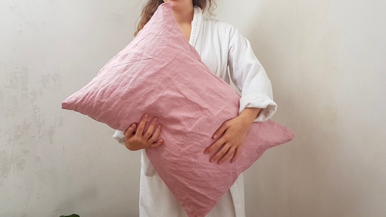 Puede incluir: Una funda de almohada de lino rosa suave sostenida por una persona que lleva una bata blanca. La funda de almohada est&aacute; hecha de una tela natural y transpirable.