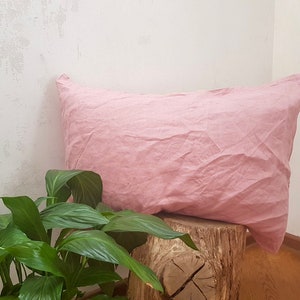 Puede incluir: Una almohada de lino rosa suave apoyada en un tronco de &aacute;rbol. La almohada est&aacute; ligeramente arrugada y tiene un aspecto natural y relajado.