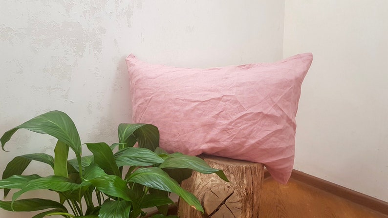 Puede incluir: Una almohada de lino rosa suave apoyada en un tronco de &aacute;rbol. La almohada est&aacute; ligeramente arrugada y tiene un aspecto natural y relajado.