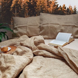 Puede incluir: Un juego de cama de lino en color beige natural con un libro y un pastel en una bandeja de madera. La ropa de cama est&aacute; dispuesta en una cama con dos almohadas. El fondo es una imagen borrosa de un bosque.