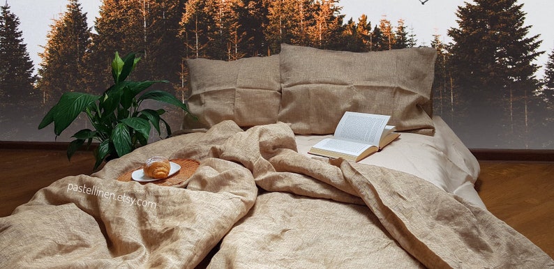 Puede incluir: Un juego de cama de lino en color beige natural con un libro y un pastel en una bandeja de madera. La ropa de cama est&aacute; dispuesta en una cama con dos almohadas. El fondo es una imagen borrosa de un bosque.