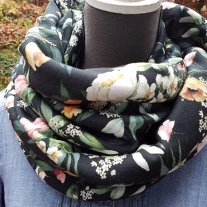 Könnte beinhalten: Schwarzer Loop-Schal mit einem Blumenmuster aus weißen, rosa und gelben Blüten und grünen Blättern. Der Schal ist um den Hals einer Schaufensterpuppe gewickelt, und der Hintergrund ist unscharf.