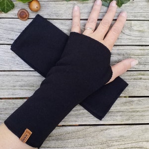 Könnte beinhalten: Schwarze fingerlose Handschuhe aus weichem Stoff. Die Handschuhe sind in der Hälfte gefaltet und haben ein kleines braunes Leder-Etikett mit dem Wort "MADE" darauf.