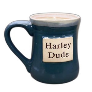 Pode incluir: Uma caneca de cerâmica azul-petróleo escuro com acabamento brilhante e borda clara. Uma placa retangular de cor branco-sujo está fixada na frente, com o texto preto em relevo Harley Dude. A caneca possui uma alça curva no lado esquerdo.