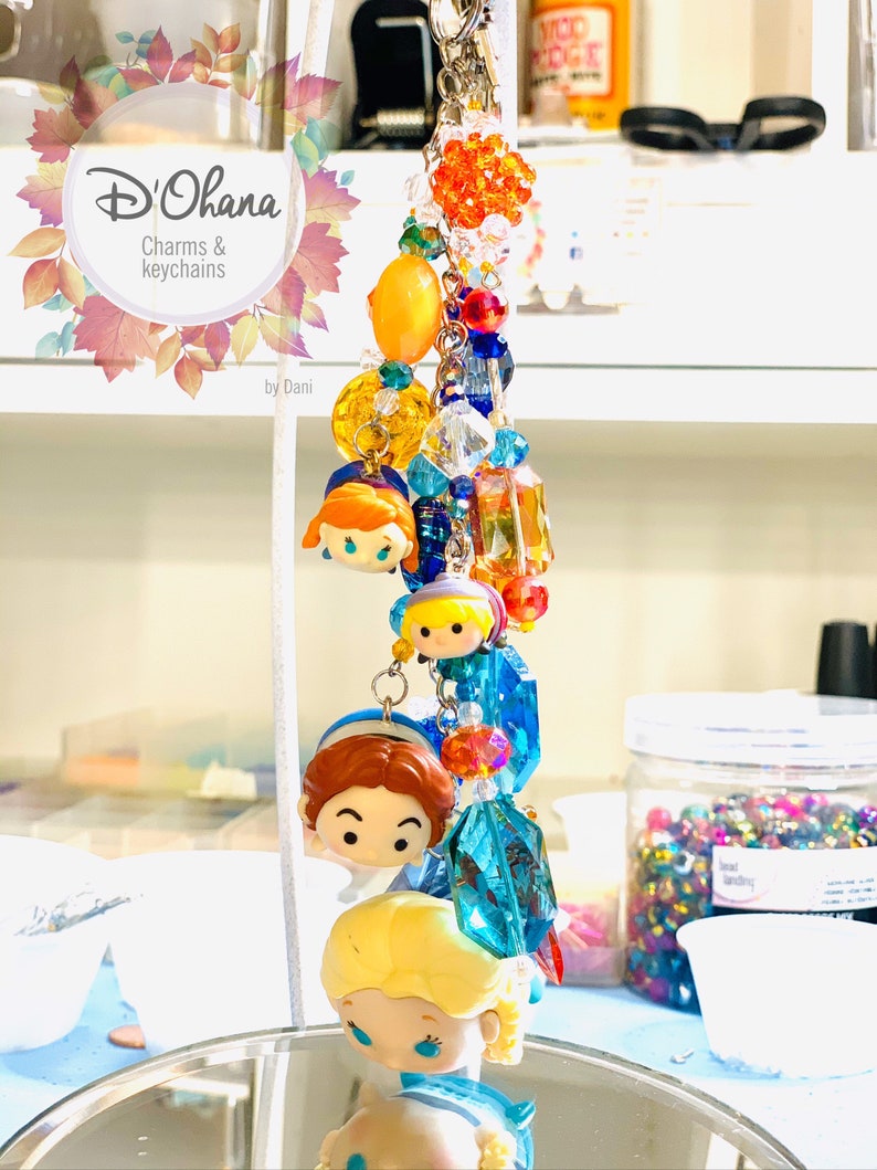 May include: A colorful charm keychain featuring Disney characters with a variety of beads, including clear, orange, yellow, blue, and red. The keychain is labeled "D'Ohana Charms & Keychains by Dani".