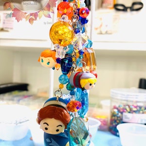 May include: A colorful charm keychain featuring a variety of beads, crystals, and small figurines. The keychain includes a blue and white figurine of Elsa and Anna from Frozen, a red and yellow figurine of a character from Tsum Tsum, and a variety of colorful beads and crystals.