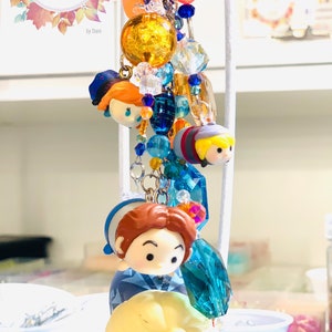 May include: A colorful charm keychain featuring characters from the animated film Frozen. The keychain includes a blue and white charm with a character from Frozen, a blue and white charm with a character from Frozen, a blue and white charm with a character from Frozen, and a blue and white charm with a character from Frozen. The keychain is adorned with blue, white, and gold beads.